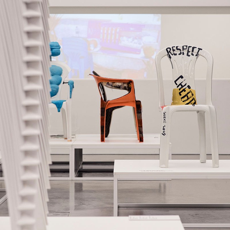 Customised monobloc chairs displayed on white plinths in a museum, one painted with the words Respect Cheap